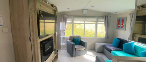 A compact living space with a flat-screen TV mounted on a wooden cabinet, a sofa set, a small table, and a window with curtains.