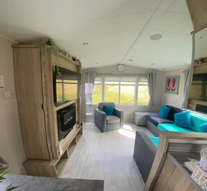 Un espace de vie compact comprenant un téléviseur à écran plat fixé à un meuble en bois, un ensemble de canapés, une petite table et une fenêtre avec des rideaux.
