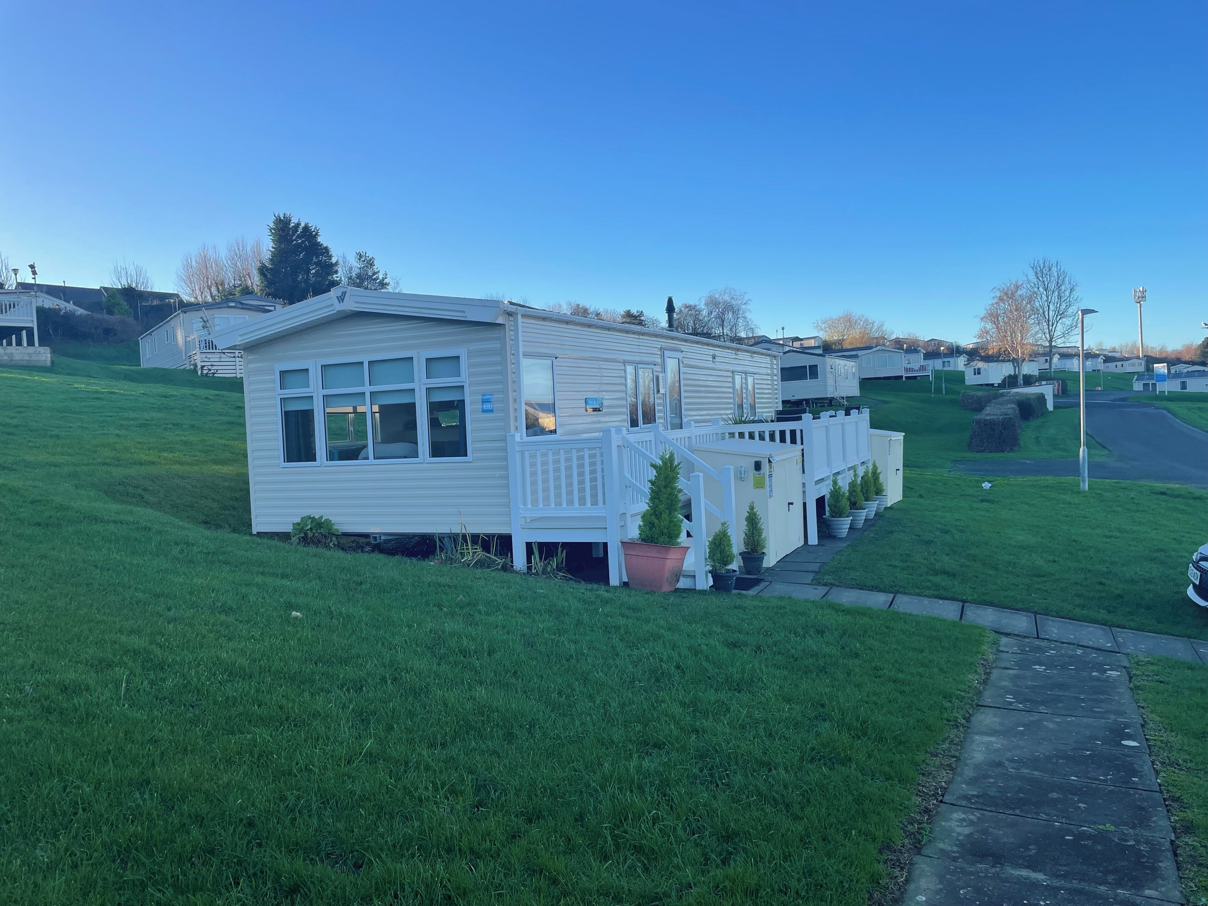 Une maison mobile de plain-pied, dotée d’une véranda clôturée, d’un petit jardin et d’une allée pavée.