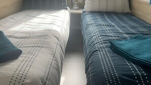 Une petite pièce, de taille modeste, comprenant deux lits, une fenêtre avec des rideaux, une table de chevet avec une lampe, et donnant sur un espace verdoyant à l’extérieur.