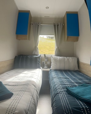 Een kleine, compacte kamer met twee bedden, een raam met gordijnen, een nachtkastje met lamp en uitzicht op een grasveld.