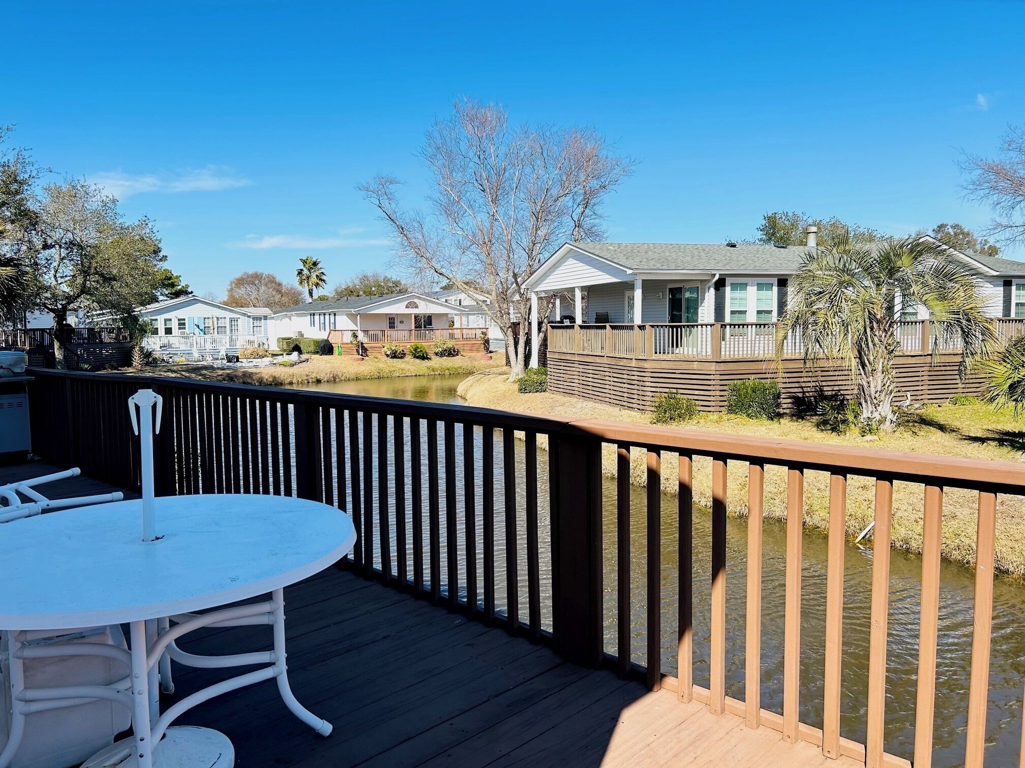 A wooden deck with a white table and chairs, overlooking a body of water and a row of houses.