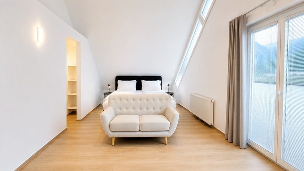 Une chambre moderne avec un grand lit, un canapé blanc et une vue sur l’extérieur, accessible par une porte-fenêtre coulissante.