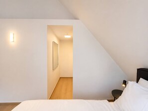 Une chambre avec un plafond en pente, un lit avec une literie blanche, une table de chevet, une lampe et un tableau encadré au mur.