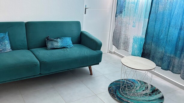 Ein blaustürkisnes Sofa mit zwei Kissen, ein kleiner runder Tisch und ein Teppich mit blau-weißem Muster, sowie eine Tür mit dem Schild 'Welcome to Paradise'.