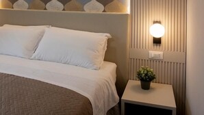A neatly made bed with a quilted cover, a wall with a geometric patterned wallpaper, a small bedside table with a potted plant, and a wall-mounted light fixture. - Economy Room from 1 to 3 persons (Roma)