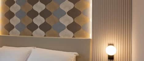 A neatly made bed with a quilted cover, a wall with a geometric patterned wallpaper, a small bedside table with a potted plant, and a wall-mounted light fixture.
