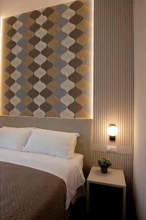 Ein ordentlich bezogenes Bett mit einer gesteppten Decke, eine Wand mit geometrischem Wandpapier, ein kleiner Nachttisch mit einem Topfblumen und eine wandmontierte Leuchte.