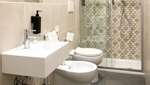 A modern bathroom with a white sink, two toilets, a glass-enclosed shower, and patterned tiles. - Economy Room from 1 to 3 persons (Roma)