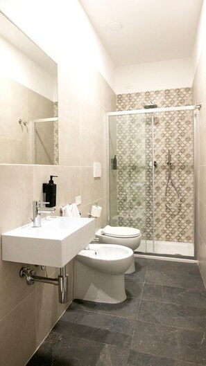 Ein modernes Badezimmer mit einem weißen Waschbecken, zwei Toiletten, einer Duschkabine aus Glas und einem Musterfliesenbelag.
