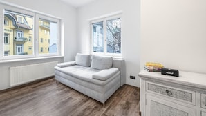 Une pièce avec un canapé gris, un meuble blanc et un radiateur. Il y a trois fenêtres donnant sur des bâtiments à l’extérieur.