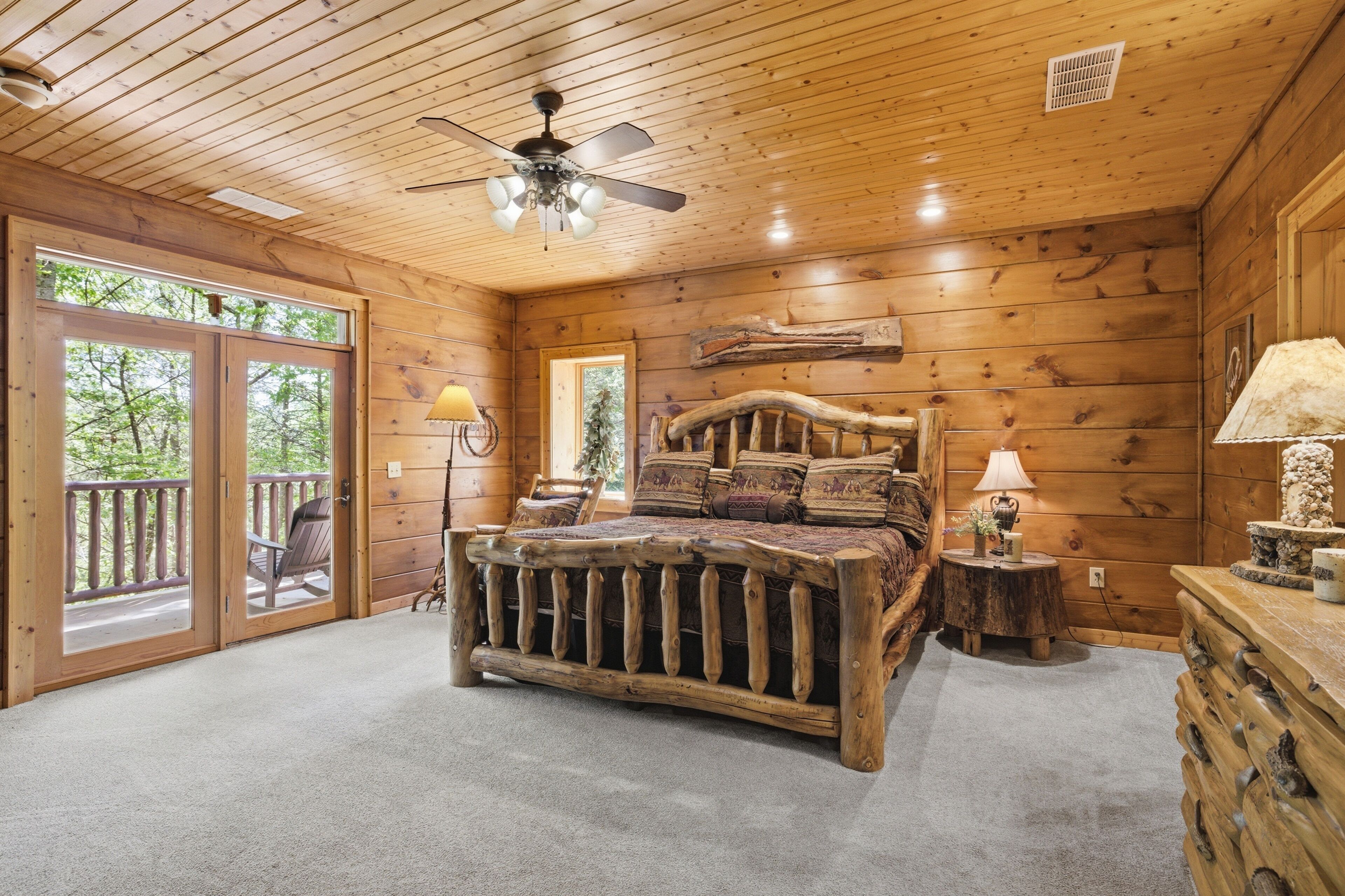 Une chambre dans un chalet en rondins, avec un grand lit, un ventilateur de plafond, une commode en bois et une vue à travers les portes vitrées.