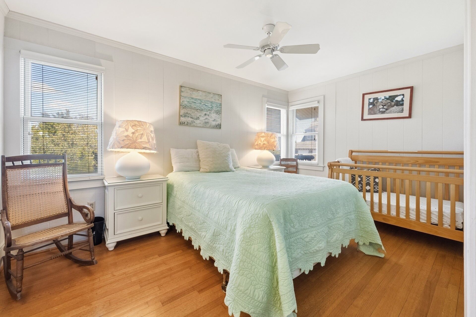 A bedroom with a bed, a wooden crib, a nightstand, a chair, and a ceiling fan.