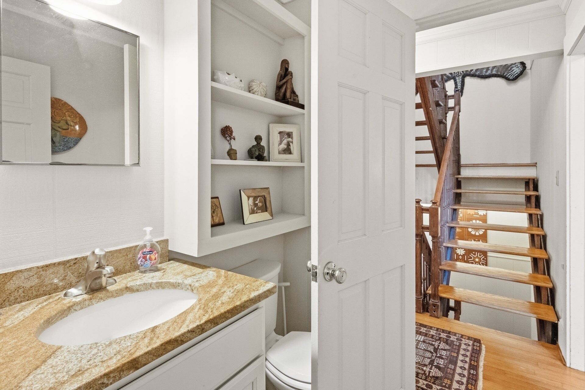 A bathroom with a granite countertop sink, a mirror, and shelves with decorative items.