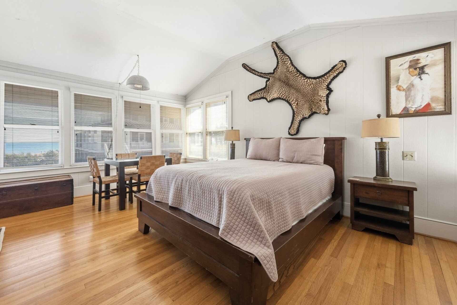 A bedroom with a large bed, a desk, and chairs. There is a window with blinds, a hanging light fixture, and a wall-mounted animal print.