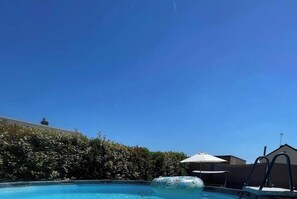 A swimming pool with a lounge chair and umbrella, surrounded by greenery and a building.