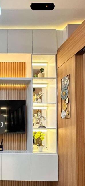 A modern living room with a flat-screen TV mounted on a white wall, wooden paneling, and built-in shelves displaying decorative items and plants. - MJM Cityscape At Red Residences Makati (Makati)