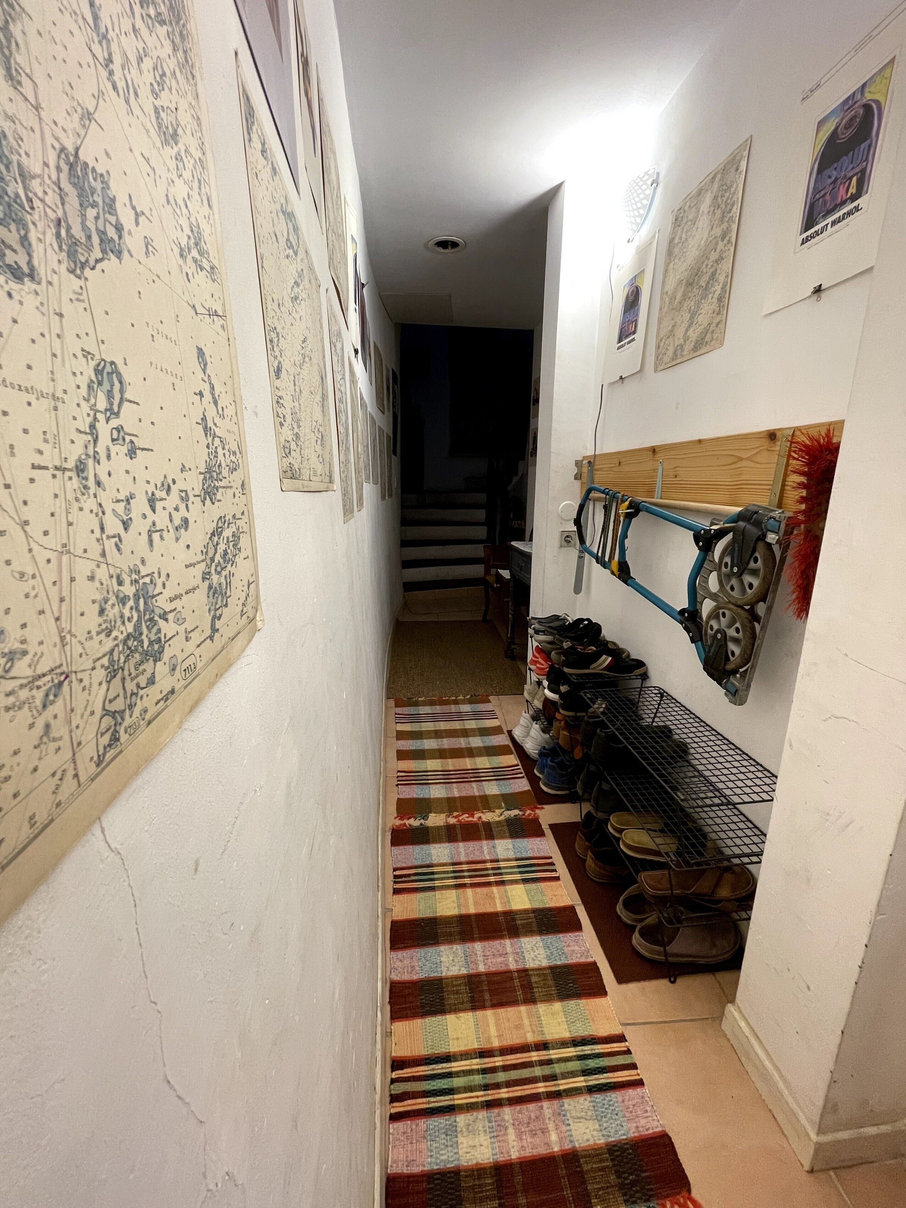 A narrow hallway with a map on the left wall, a shoe rack on the right, and a staircase in the background.