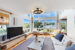 A spacious living room with a large TV, comfortable seating, and large windows offering a view of the outdoors. - Dog-friendly riverfront home with dock, 2 balconies, waterside patio, & grill (Annapolis)