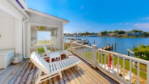 Una terraza de madera con sillas y una mesa blancos, con vista a un puerto deportivo con barcos y casas al fondo.