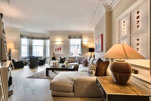 A modern living room with a sofa, coffee table, and lamps. There are windows with curtains, a fireplace, and a wall with decorative panels. - 3-bed Knightsbridge Harrods View Best Location (London)