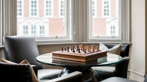 Un échiquier posé sur une table en verre, dans une pièce où se trouvent des fauteuils en cuir et de grandes fenêtres donnant sur des immeubles.