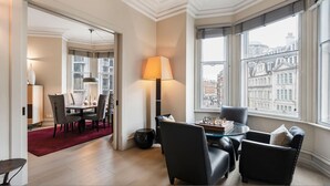 Une chambre d’hôtel moderne dotée d’un coin repas, d’une grande fenêtre offrant une vue sur la ville et d’un espace salon confortable avec un jeu d’échecs.