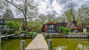 Eine Holzstege führt zu einem Haus mit Balkon, umgeben von Bäumen und Wasser.