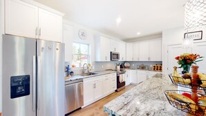 Una cocina moderna con armarios blancos, electrodomésticos de acero inoxidable, encimera de mármol y una isla central con una cesta de frutas.
