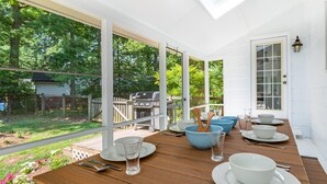 Ein überdachter Essbereich im Freien mit einem Holztisch, gedeckt für vier Personen, umgeben von großen Fenstern mit Blick auf den Garten.
