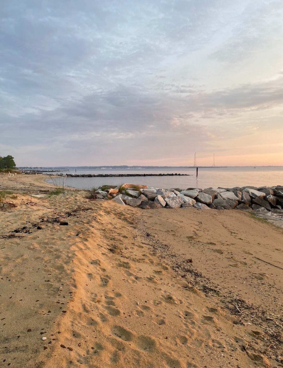 Ein Strand mit einem steinernen Wellenbrecher, sandigem Ufer und einem weiter entfernten Steg.