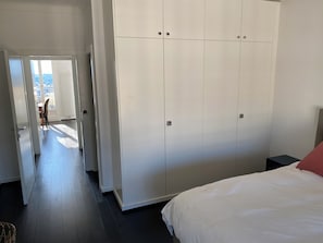 Un dormitorio con un armario grande, una cama con ropa de cama blanca y vistas a través de una puerta abierta.