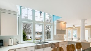 A modern kitchen with large windows, a central island, and white cabinetry. - Beautiful waterfront home on The Chesapeake Bay with private dock & beach access (Annapolis)