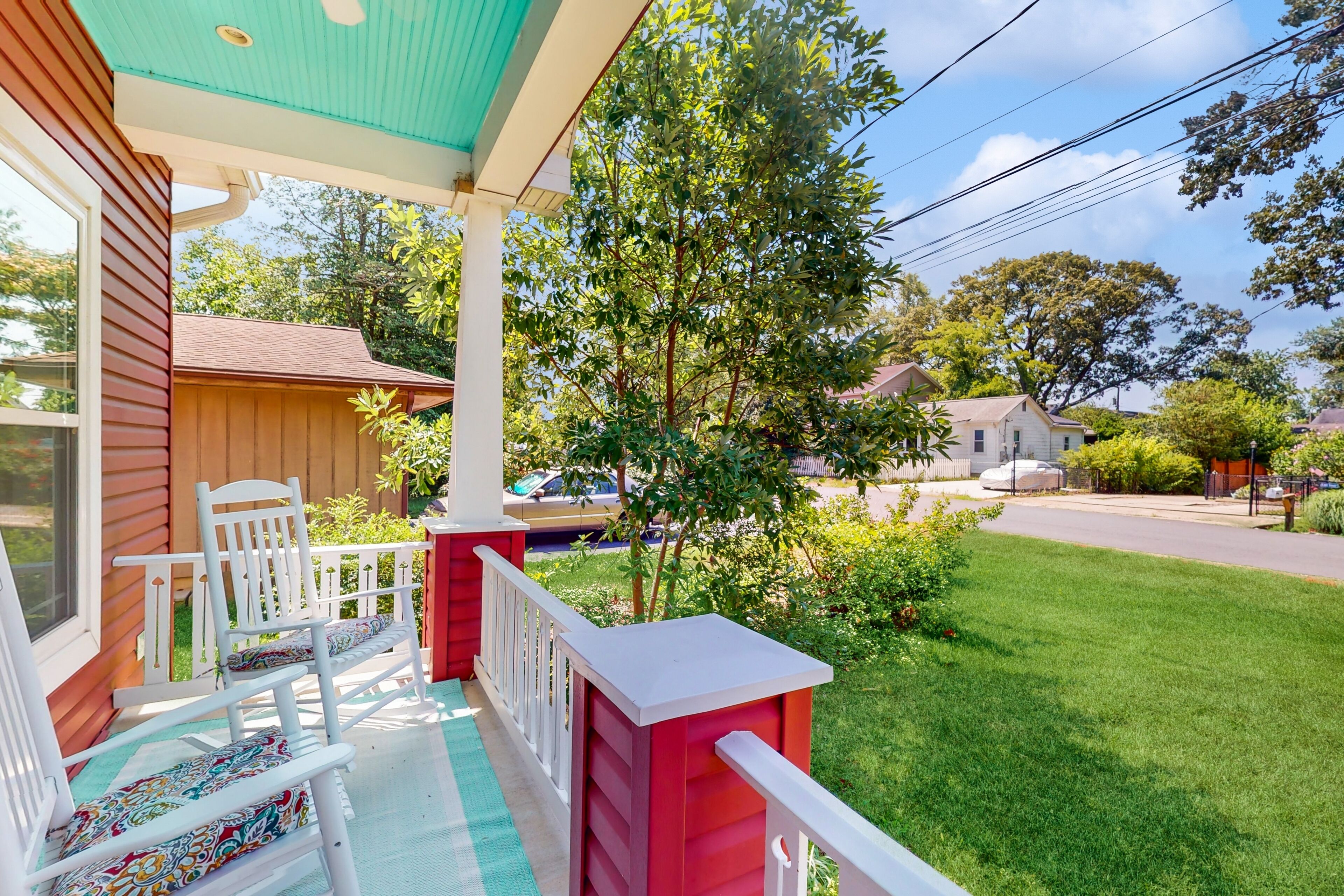 A porch with two white rocking chairs and a red railing, overlooking a green lawn and neighboring houses.
