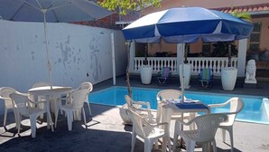 A poolside area with white plastic chairs and tables, a blue and white umbrella, and a white fence with decorative elements. - House in Rifaina, with swimming pool, close to the beach. (Rifaina)