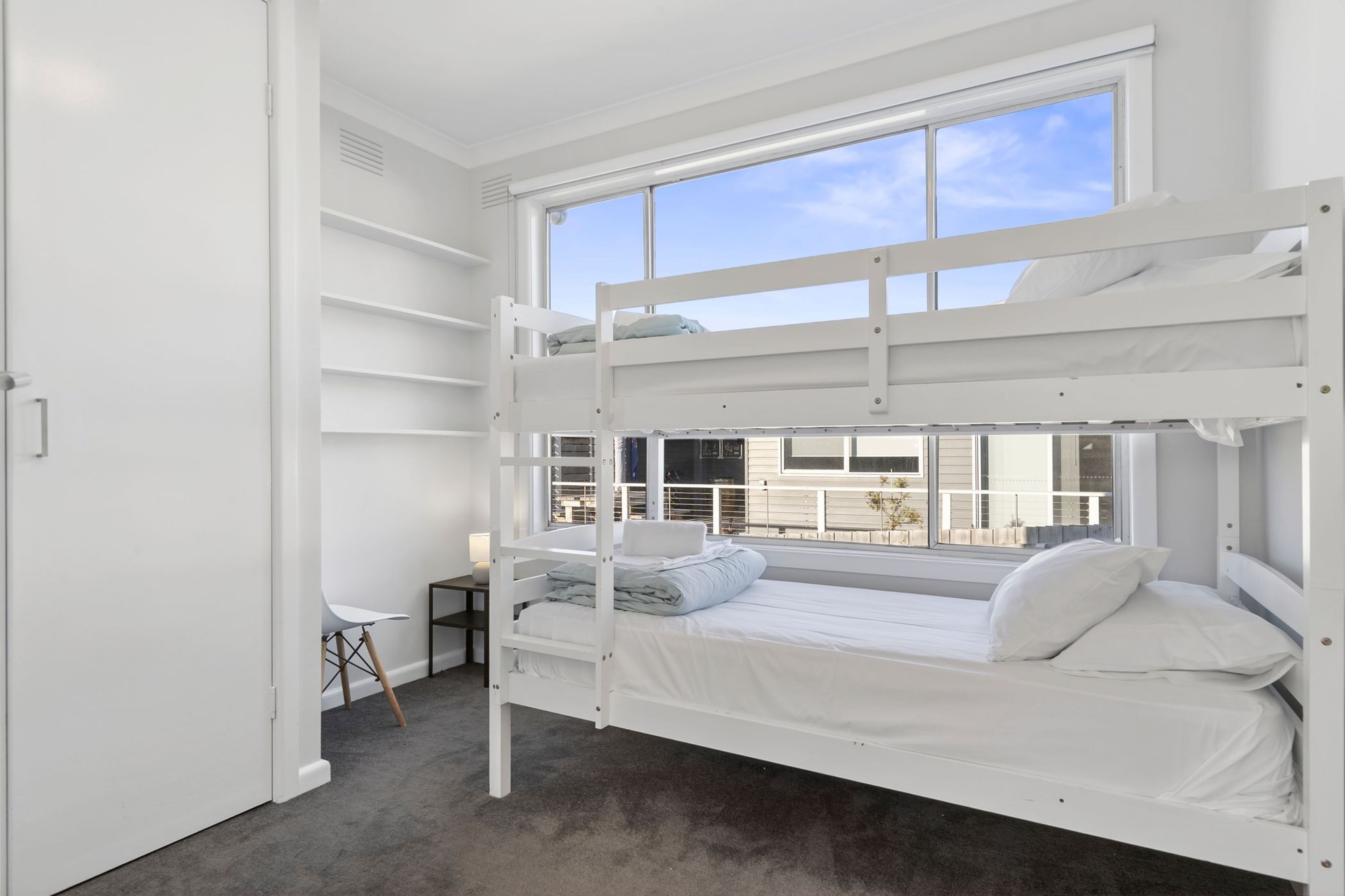 A bunk bed room with a desk and chair, a window with a view of buildings, and a carpeted floor.