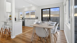 A modern kitchen with white cabinets, a dining area with a wooden table, and a view of a residential area through large windows. - Days by the Bay (Apollo Bay)