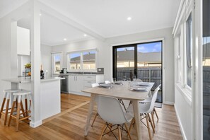 Eine moderne Küche mit weißen Schränken, ein Essbereich mit einem Holztisch und Blick auf eine Wohngegend durch große Fenster.