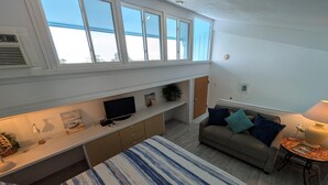 Quarto espaçoso com uma cama grande, um sofá, uma TV e uma mesa com um computador. Há janelas amplas e vista do céu.