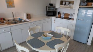 Cozinha com armários brancos, pia, fogão e geladeira. Há uma mesa de jantar com cadeiras e uma tigela sobre ela.