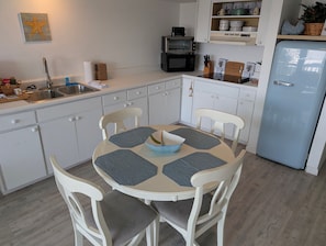 A kitchen with white cabinets, a sink, a stove, and a refrigerator. There is a dining table with chairs and a bowl on it. - Park Place -Parkside Dream -Downtown w/ Gulf View! (Cedar Key)
