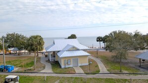 A small, single-story building with a metal roof, situated on a grassy area with a view of the ocean. - Park Place -Parkside Dream -Downtown w/ Gulf View! (Cedar Key)