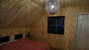 Ein Zimmer mit geneigter Decke, einem Bett mit rot kariertem Überwurf, einem kleinen Tisch mit einer Laterne und einem Fenster mit Blick auf den Nachthimmel.