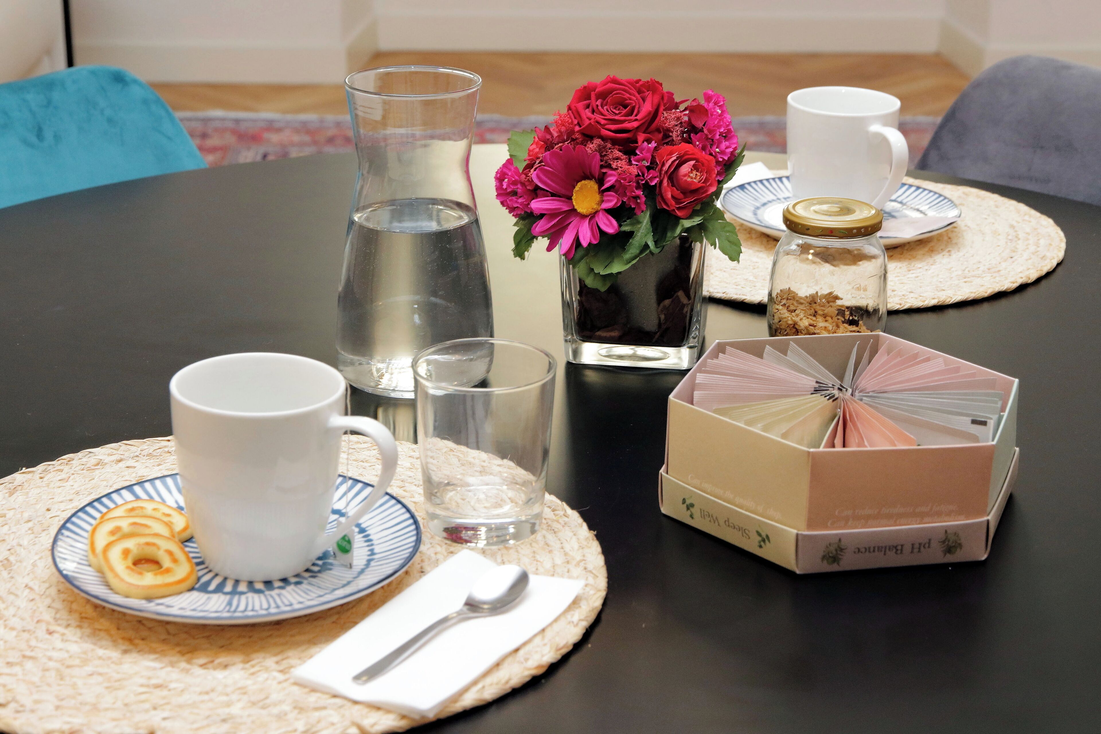 Een gedekte tafel met een witte mok, een glas water, een bord met koekjes, een kleine vaas met bloemen, een suikerpot en een theedoos.