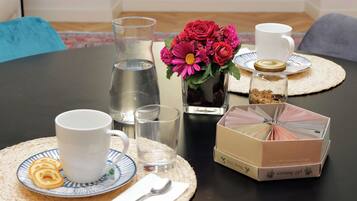 Een gedekte tafel met een witte mok, een glas water, een bord met koekjes, een kleine vaas met bloemen, een suikerpot en een theedoos.