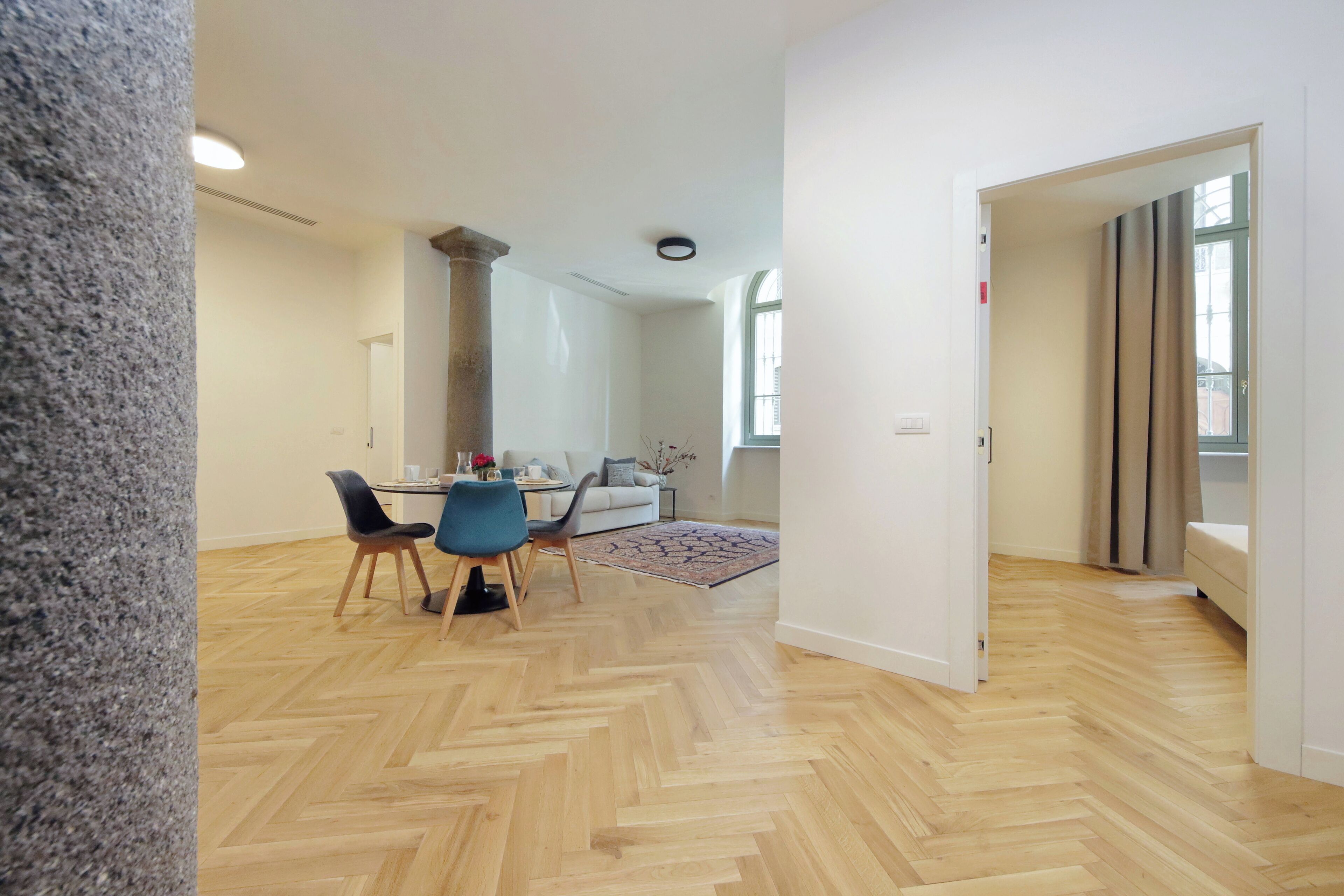 Ein geräumiges Zimmer mit Fischgrätparkett, einem Essbereich mit Tisch und Stühlen sowie einem großen Fenster mit Vorhängen.