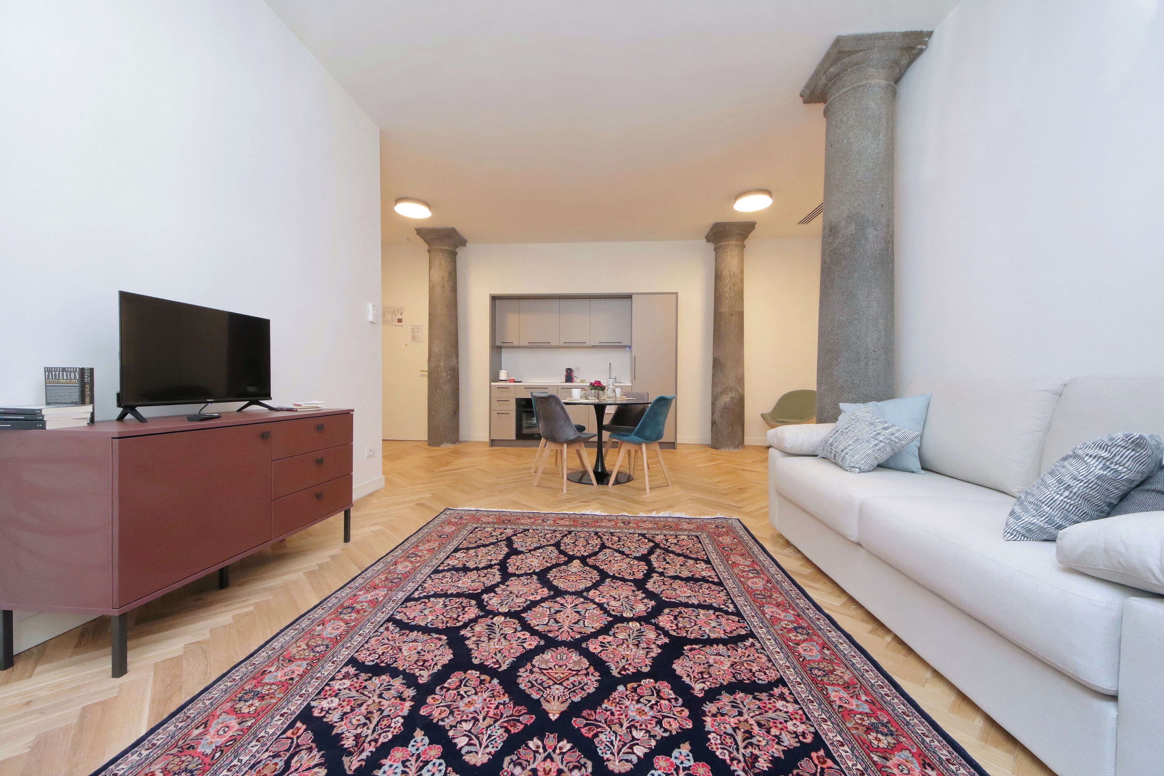 Una sala de estar con sofá, televisor sobre un mueble, zona de comedor con sillas y cocina al fondo.