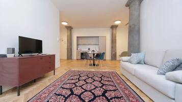 Una sala de estar con sofá, televisor sobre un mueble, zona de comedor con sillas y cocina al fondo.