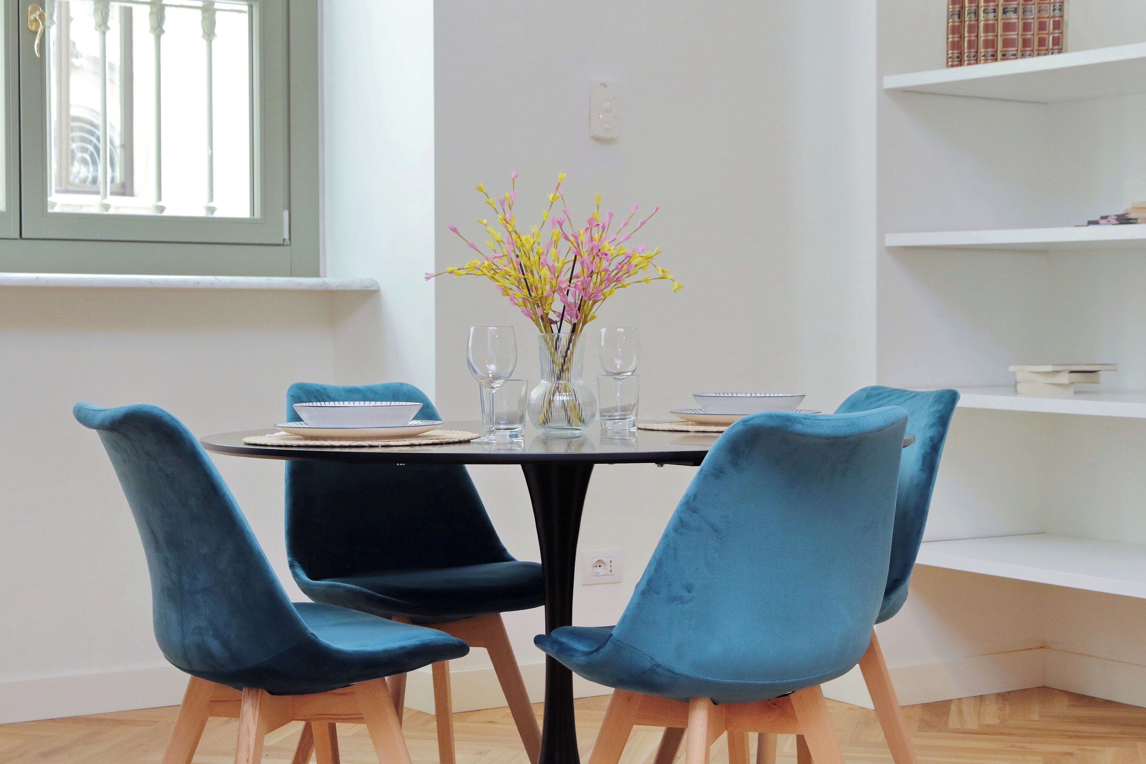 Une table à manger avec quatre chaises bleues, un vase rempli de fleurs et une fenêtre avec des barreaux.