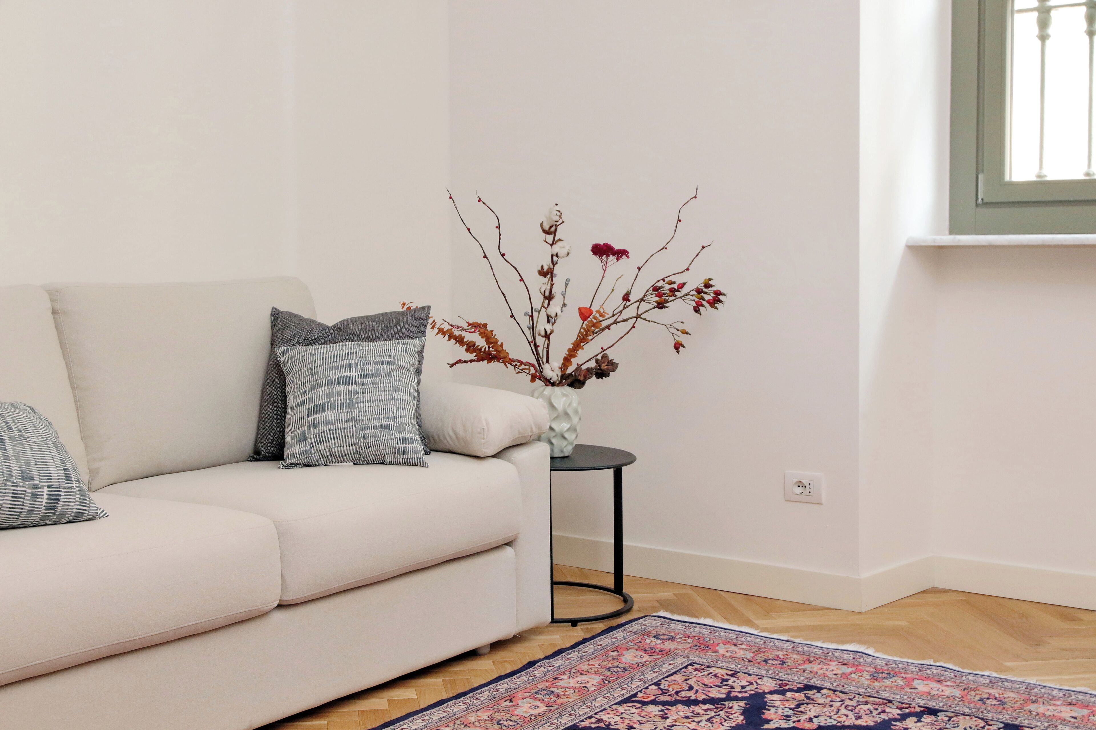 Ein weißes Sofa mit gemusterten Kissen, ein kleiner Beistelltisch mit einer Vase gefüllt mit getrockneten Ästen und ein gemusterter Teppich auf Holzdielen.